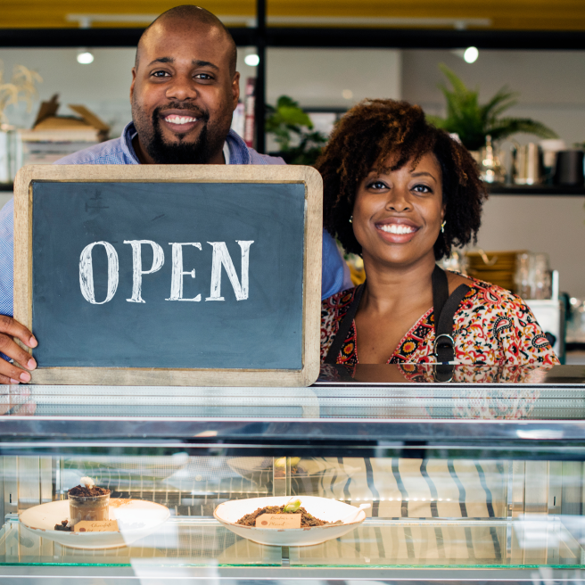 Black man and woman stand with an open sign in from of them, inside their bakery