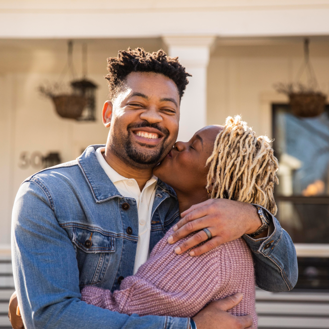 Black woman with blonde locs kisses black man in front of their house